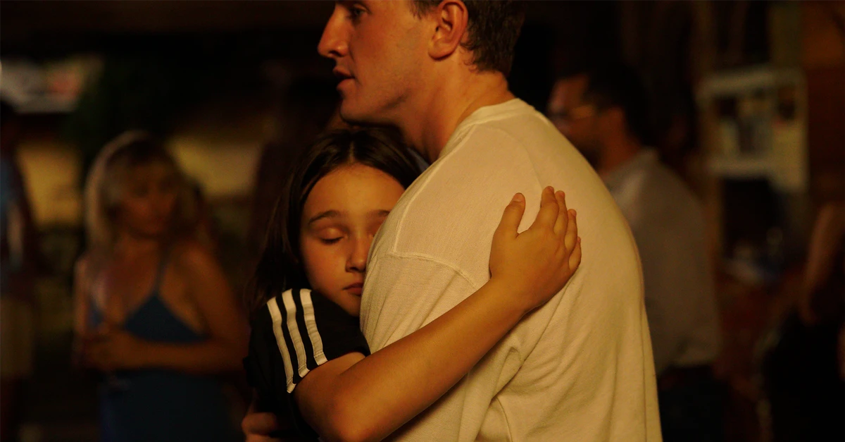 Cena do filme Aftersun, mostra pai e filha dançando abraçados. Ao fundo, vemos pessoas desfocadas. 