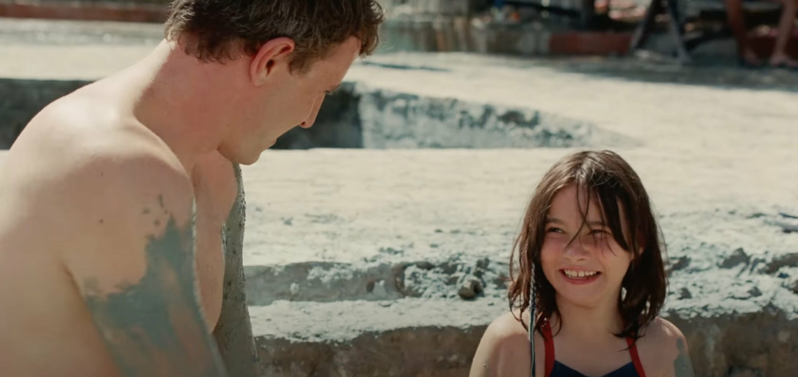 Cena do filme Aftersun, mostra pai e filha numa piscina de lama, se sujando. Eles sorriem um para o outro e está de dia e ensolarado. 