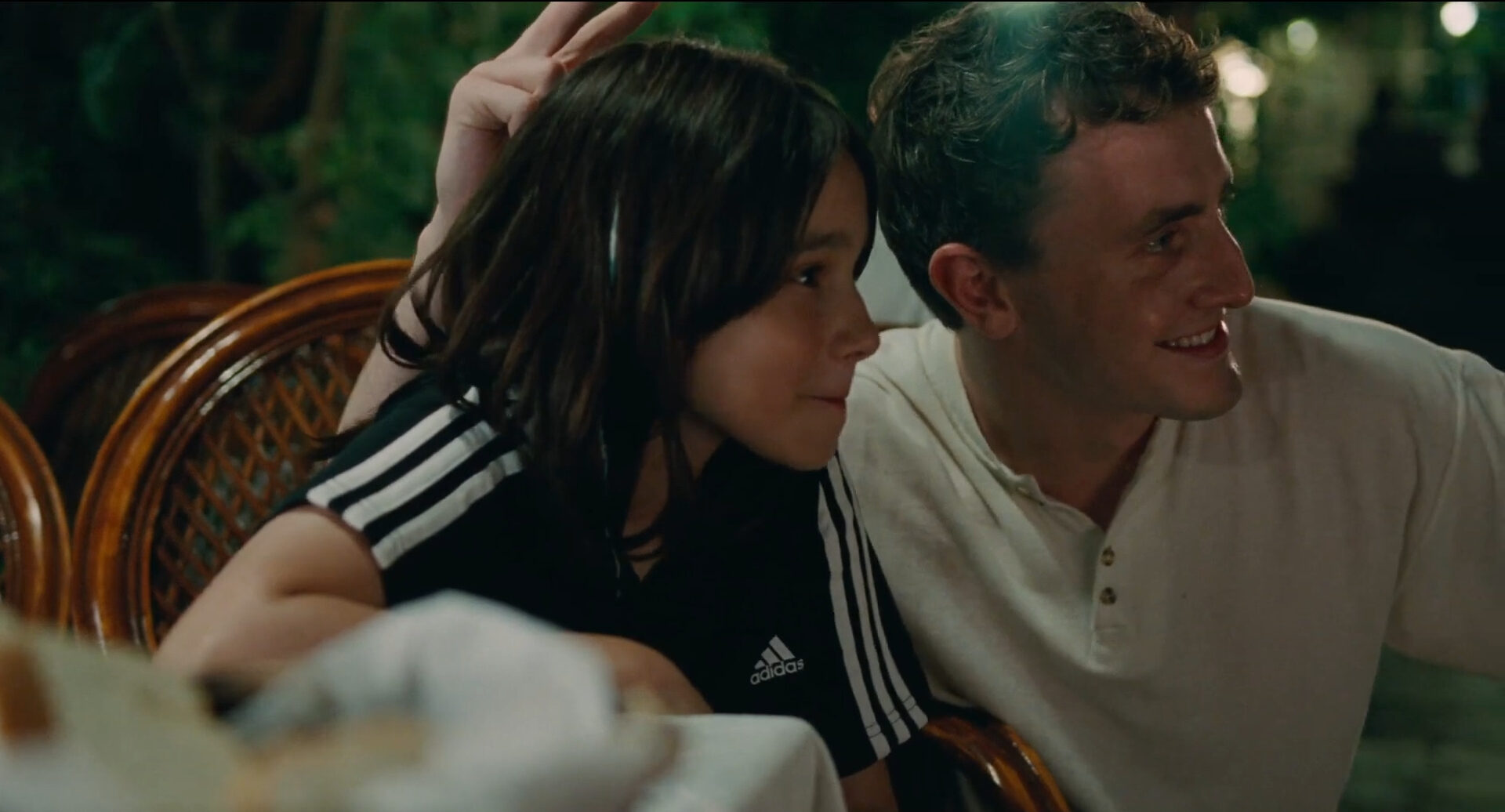 Cena do filme Aftersun, mostra pai e filha posando para uma foto, sorrindo. Por trás da menina, ele faz “chifrinho” com a mão na cabeça dela.