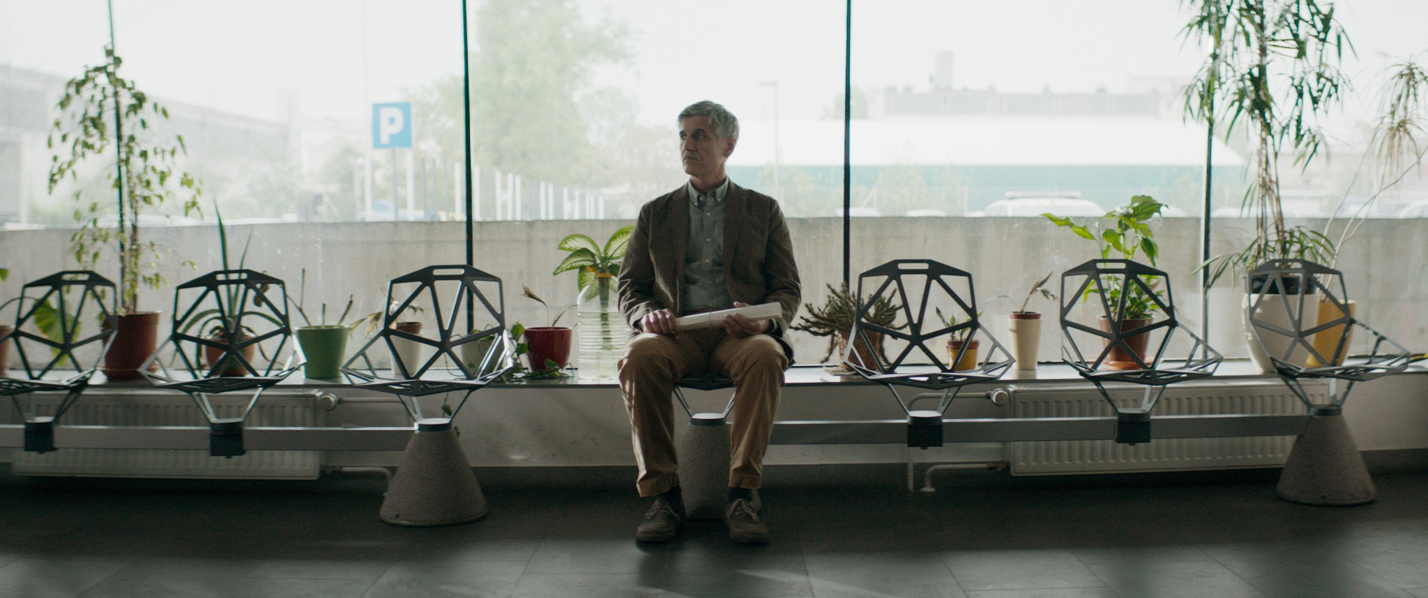 Cena do filme Inventário exibe um homem de meia idade parado em uma sala de espera. Ele segura um rolo de papel no colo. À direita e no fundo, vemos algumas plantas que servem de decoração para o local. Ao fundo, vemos a cidade, embaçada pelo vidro. 