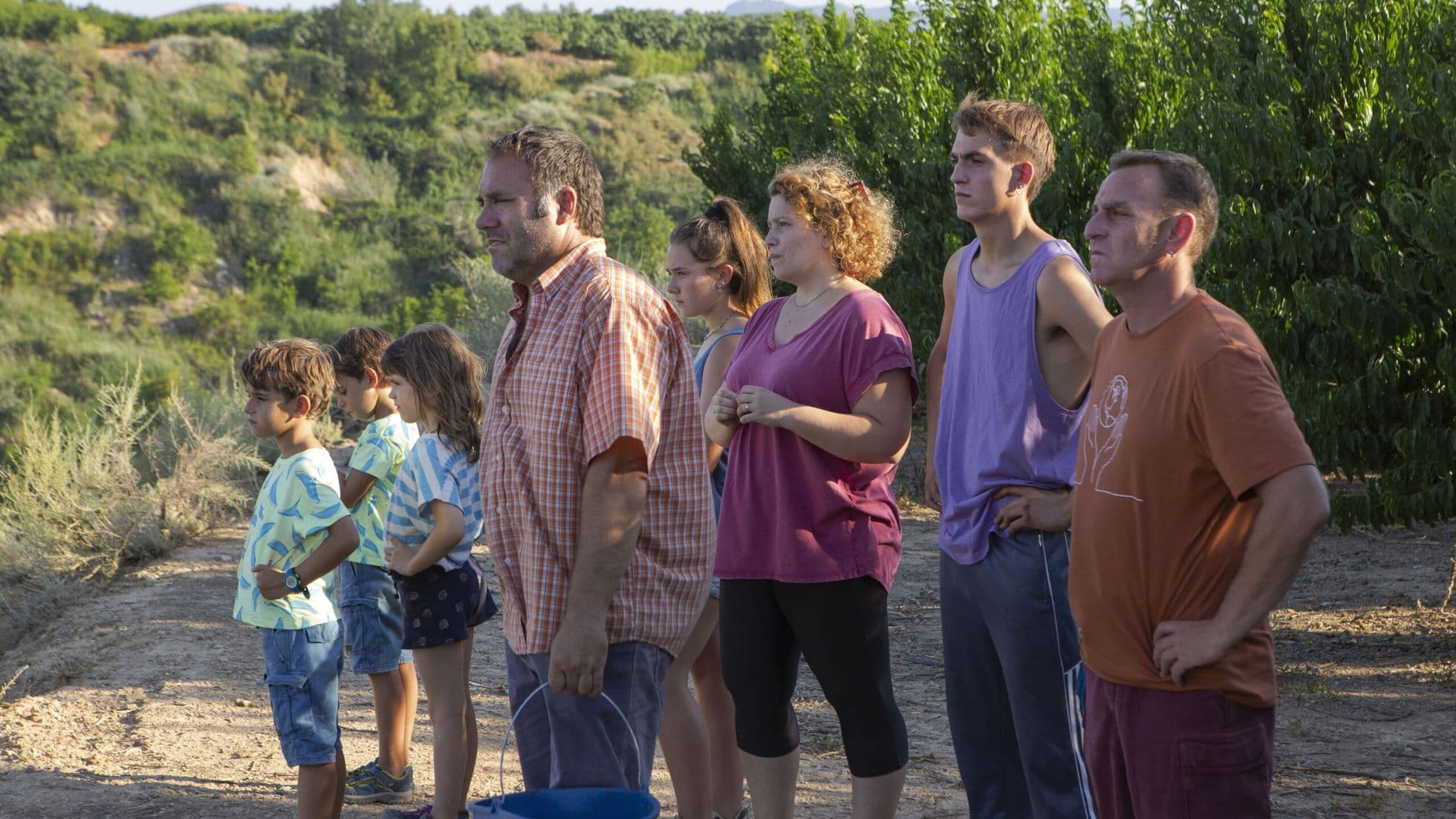 Imagem retangular que mostra uma cena do filme Algarràs. Uma família branca está em pé, todos virados de lado, olhando na direção esquerda da imagem. O fundo tem montanhas e plantações verdes.