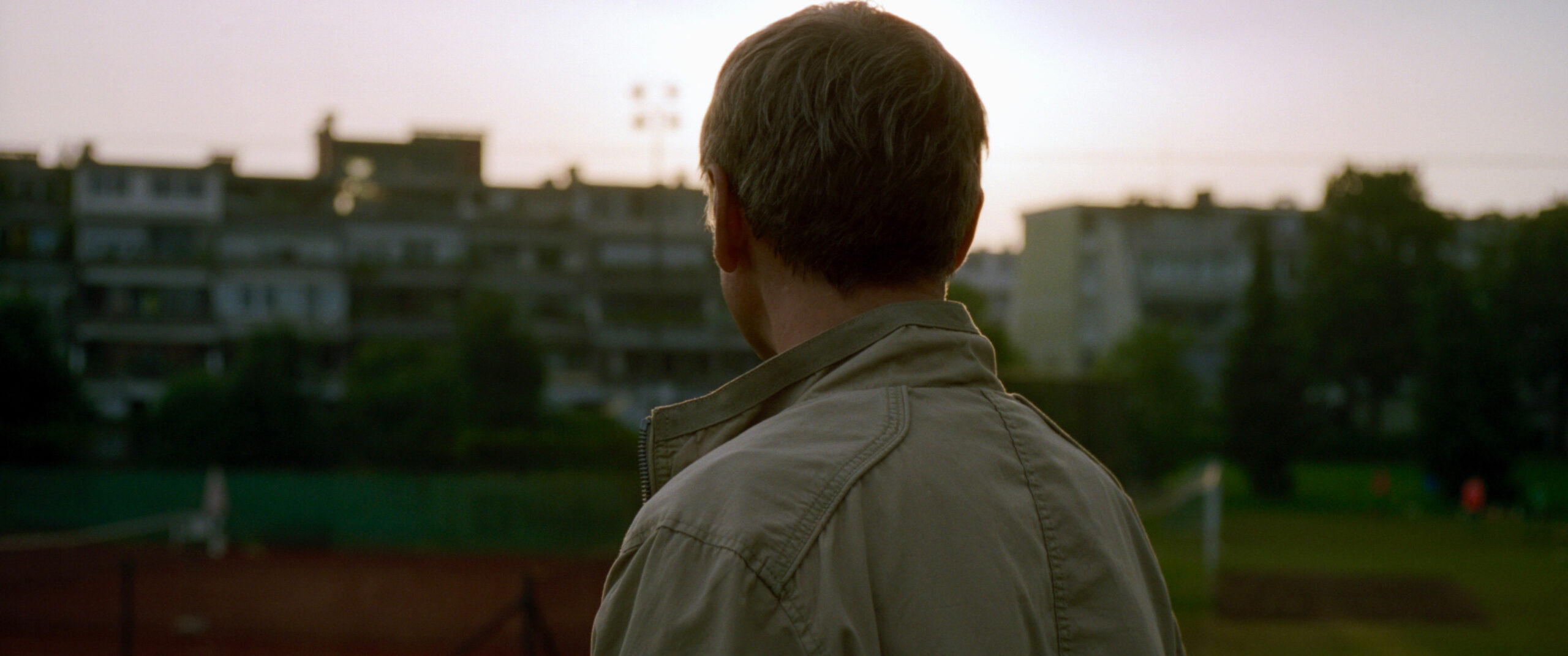 Cena do filme Inventário exibe a nuca de um homem branco que olha para apartamentos ao pôr do sol, que estão em um fundo desfocado. Ele veste uma jaqueta e tem cabelo grisalho. 