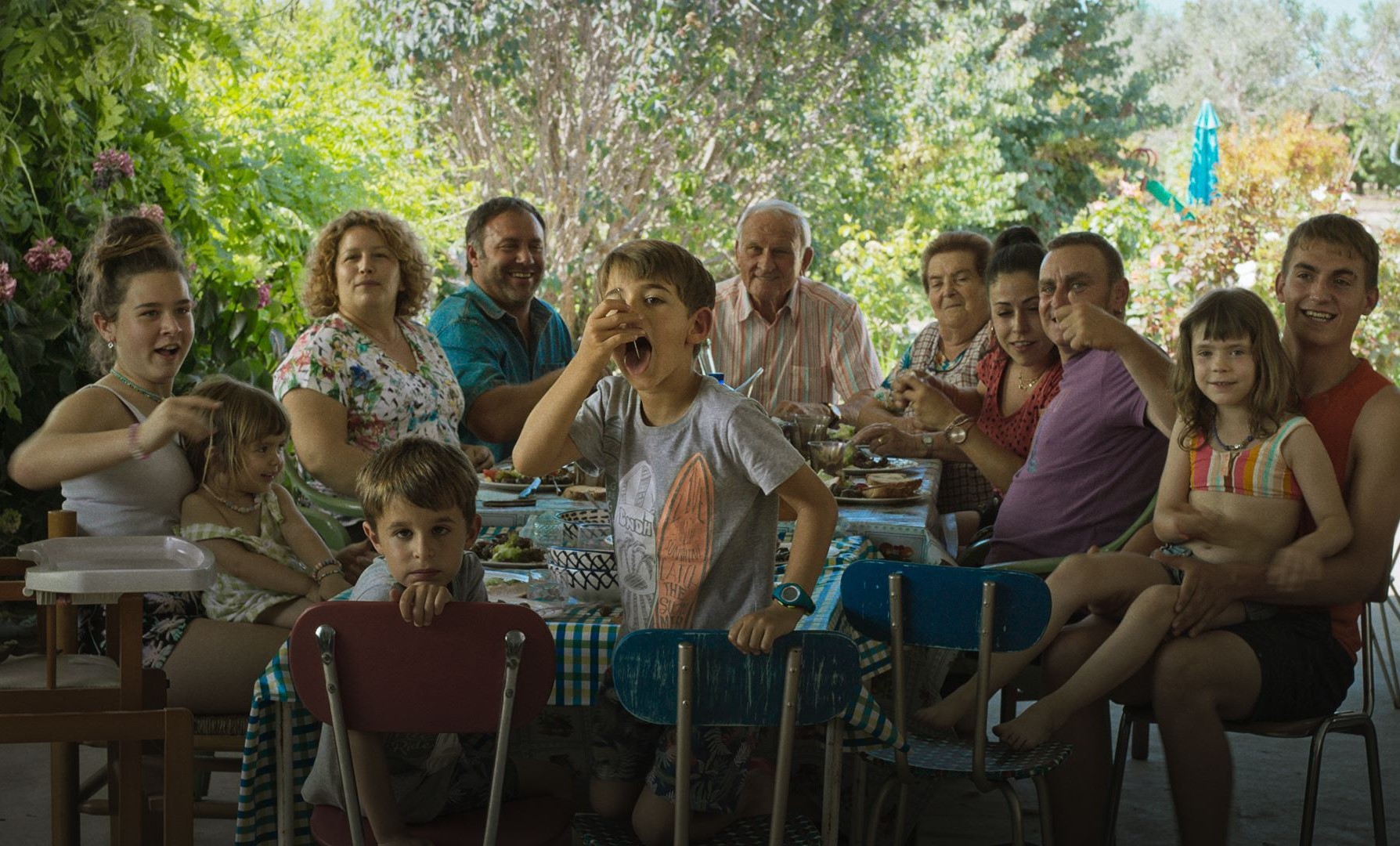 Imagem retangular que mostra uma cena do filme Algarràs. Uma família branca está reunida, sentada em uma mesa repleta de comidas. Eles estão virados para a direção da foto. Todos sorriem, se movimentam ou conversam uns com os outros.