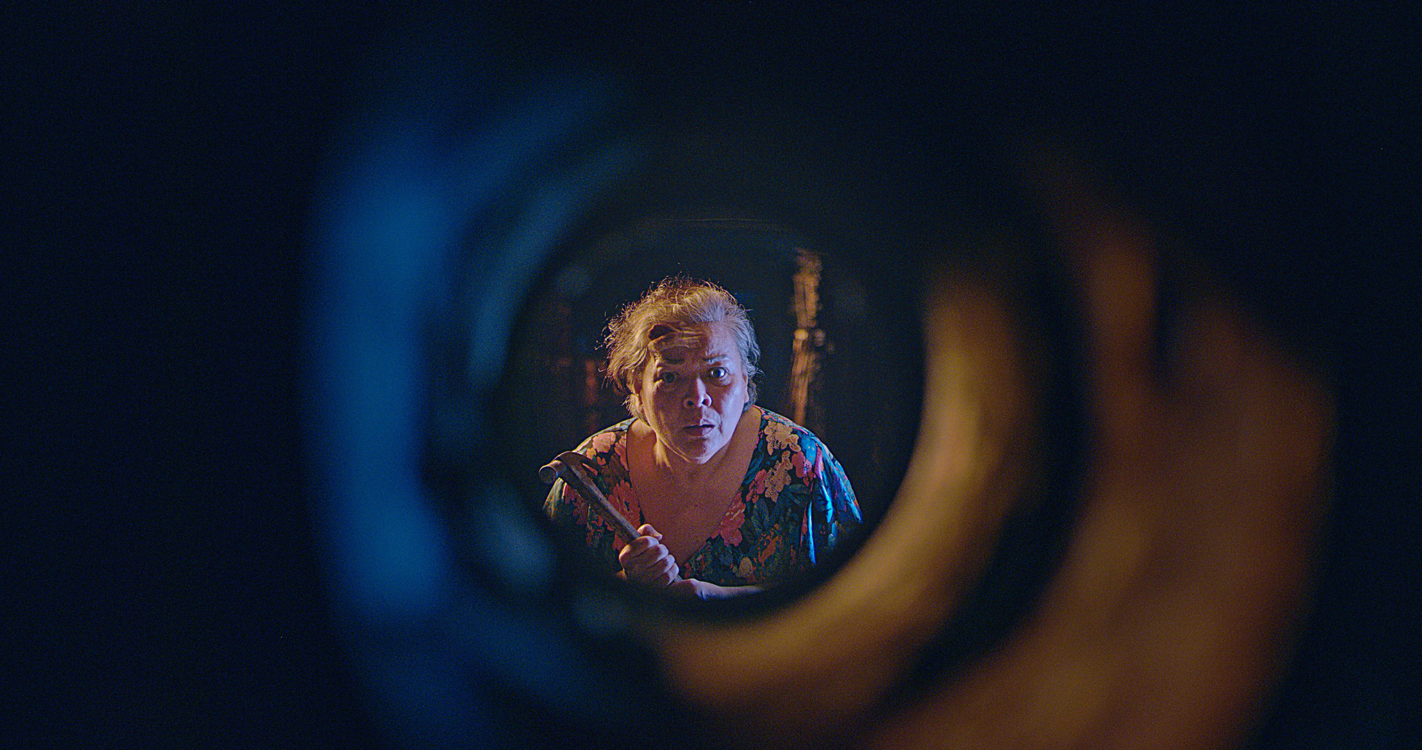 ena do filme Leonor Jamais Morrerá mostra uma idosa filipina com os olhos arregalados e segurando um martelo nas mãos. Ela olha para algo dentro de uma abertura circular, que funciona como uma moldura para sua imagem na cena. 