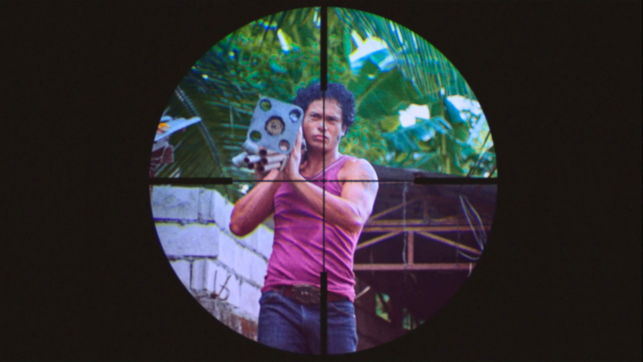 Cena do filme Leonor Jamais Morrerá mostra um homem filipino carregando canos no ombro direito. Ele veste uma regata rosa com calça jeans e tem cabelo preto. Na imagem, vemos a mira de uma arma em cima dele. 