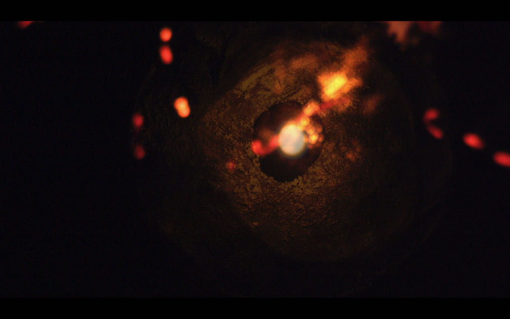 Cena do filme Objetos de Luz. Na imagem, pequenas partículas de brilho em tons de vermelho e laranja passam por um buraco escuro.