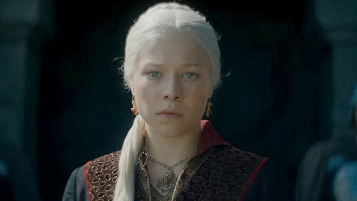 Cena de House of The Dragon. Nela vemos a personagem Rhaenyra Targaryen. Uma mulher branca, de olhos azuis e cabelos platinados. Ela usa uma roupa típica medieval preta com detalhes em vermelho. Ela também usa um colar com um dragão de três cabeças e brincos de rubi. Ao fundo, temos uma fortaleza feita de pedras