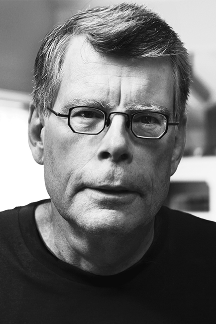  Foto do autor Stephen King. A imagem é em preto e branco. King é um homem branco de cabelos curtos. Seu rosto apresenta algumas rugas e linhas de expressão. Ele usa um óculos preto e veste uma camiseta também preta.