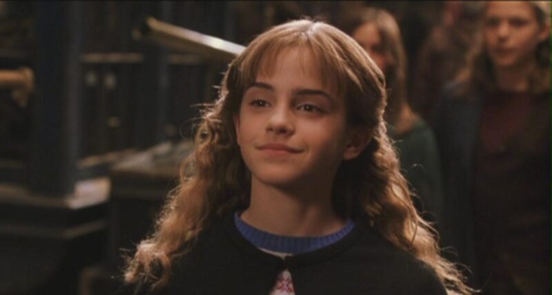 Cena do filme Harry Potter e a Câmara Secreta. O fundo da foto é desfocado com pessoas passando. No centro, está  Hermione Granger (Emma Watson), jovem branca, com cabelos castanhos ondulados, dando um leve sorriso com os lábios. Ela veste uma capa preta sobre uma blusa de gola azul.