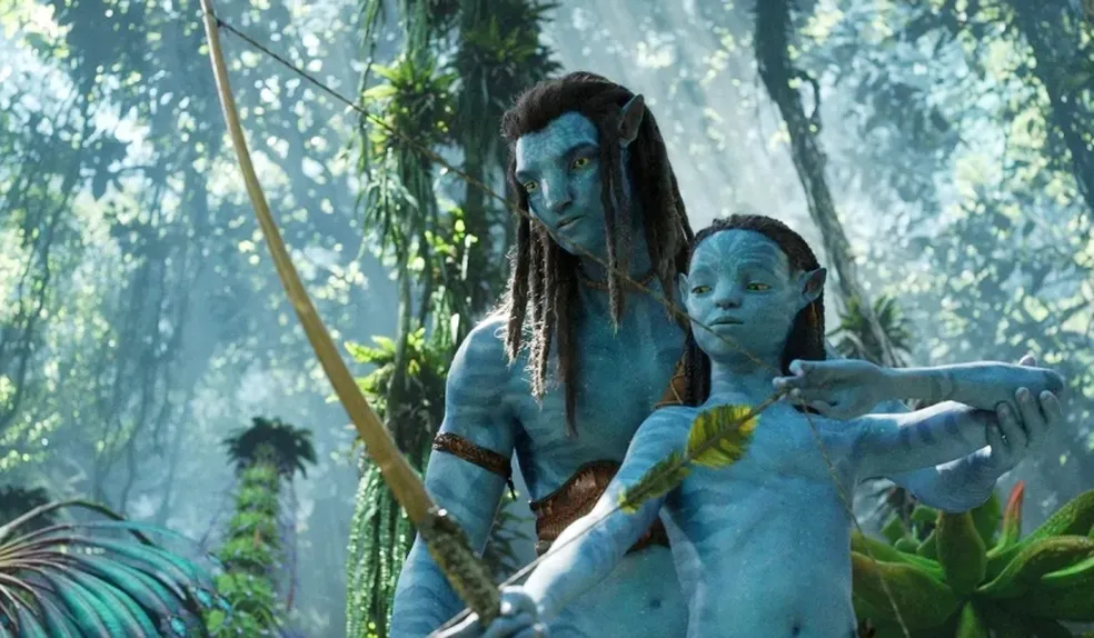 Cena do filme Avatar 2: O Caminho da Água. Nela estão dois Na’vi em meio a uma floresta. Jake Sully e seu filho. Seus olhos são grandes e amarelos, e uma estrutura corporal que equivale a dois humanos com uma cauda longa e fina. Seu corpo é coberto por uma pele azul brilhante com listras mais escuras, e seu cabelo é longo e preto.
