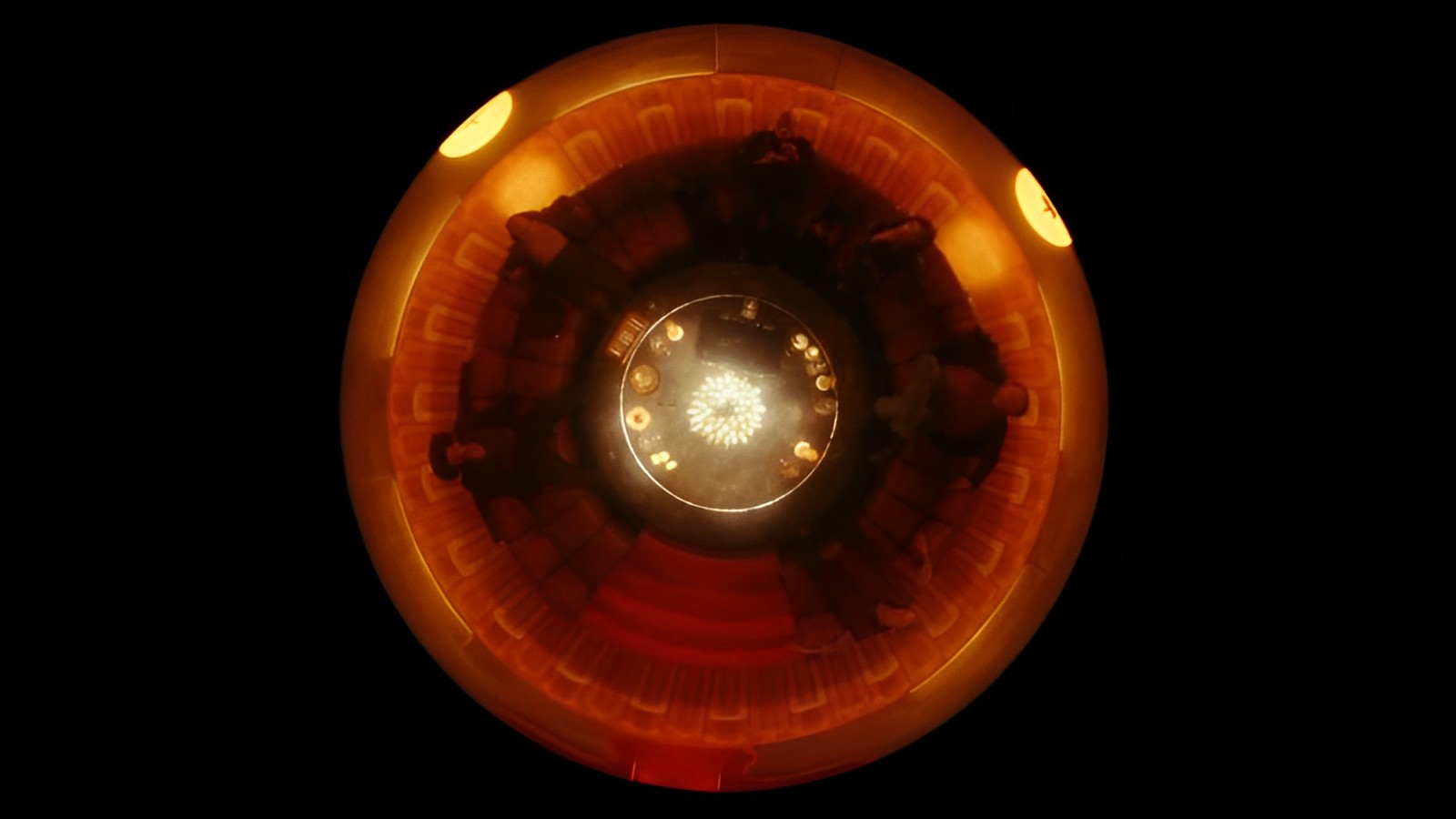 Cena da série O Gabinete de Curiosidades de Guillermo del Toro. Nela, no centro, há uma cápsula alaranjada circular apresentada de cima, como em uma visão áerea. Cinco pessoas estão espalhadas na borda da cápsula, enquanto, no centro dela, está uma mesa circular repleta de focos de luz. 