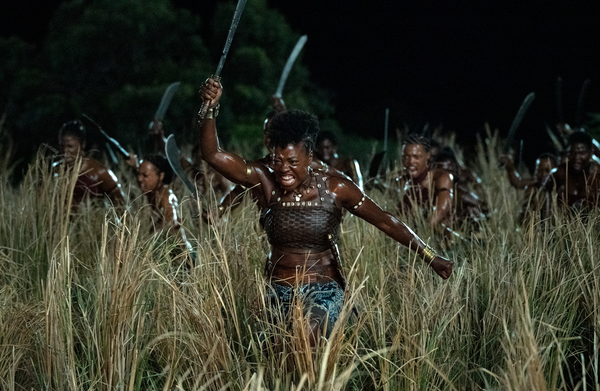 Cena do filme A Mulher Rei (2022), Nanisca (Viola Davis), vestida com um top e uma saia em tons de verde, sai com seu grupo de guerreiras de um matagal. Estão todas armadas de facões e adornadas com colares e braceletes típicos