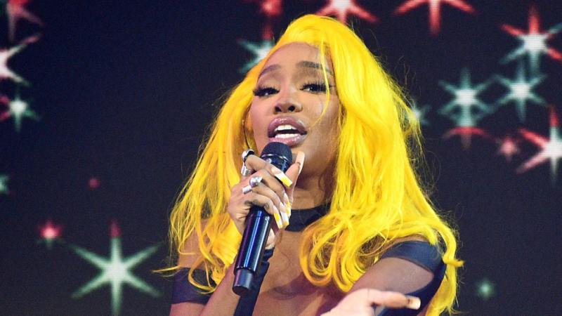 Foto da Cantora SZA, uma mulher negra, jovem com cabelos amarelos, lisos e longos. Ela está cantando com um microfone em sua mão esquerda e estendendo sua mão direita aberta ao público. Ao fundo, um telão de um palco com estrelas verdes e vermelhas.