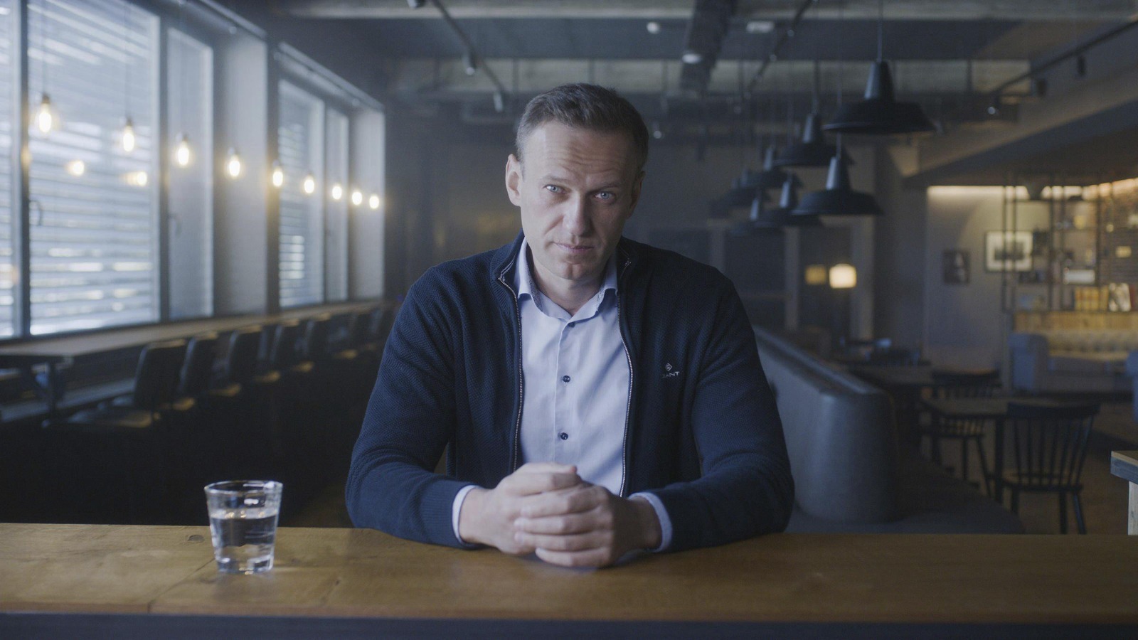 Cena do documentário Navalny. Na cena, vemos, do abdômen para cima, Alexey Navalny, um homem branco, aparentando ter cerca de 40 anos, de cabelos castanhos curtos, vestindo uma camisa azul clara e uma jaqueta azul escura, sentado por detrás de uma mesa de madeira, do que aparenta ser um bar. Ao lado esquerdo dele, vemos um copo d’água. Ele encara a câmera.