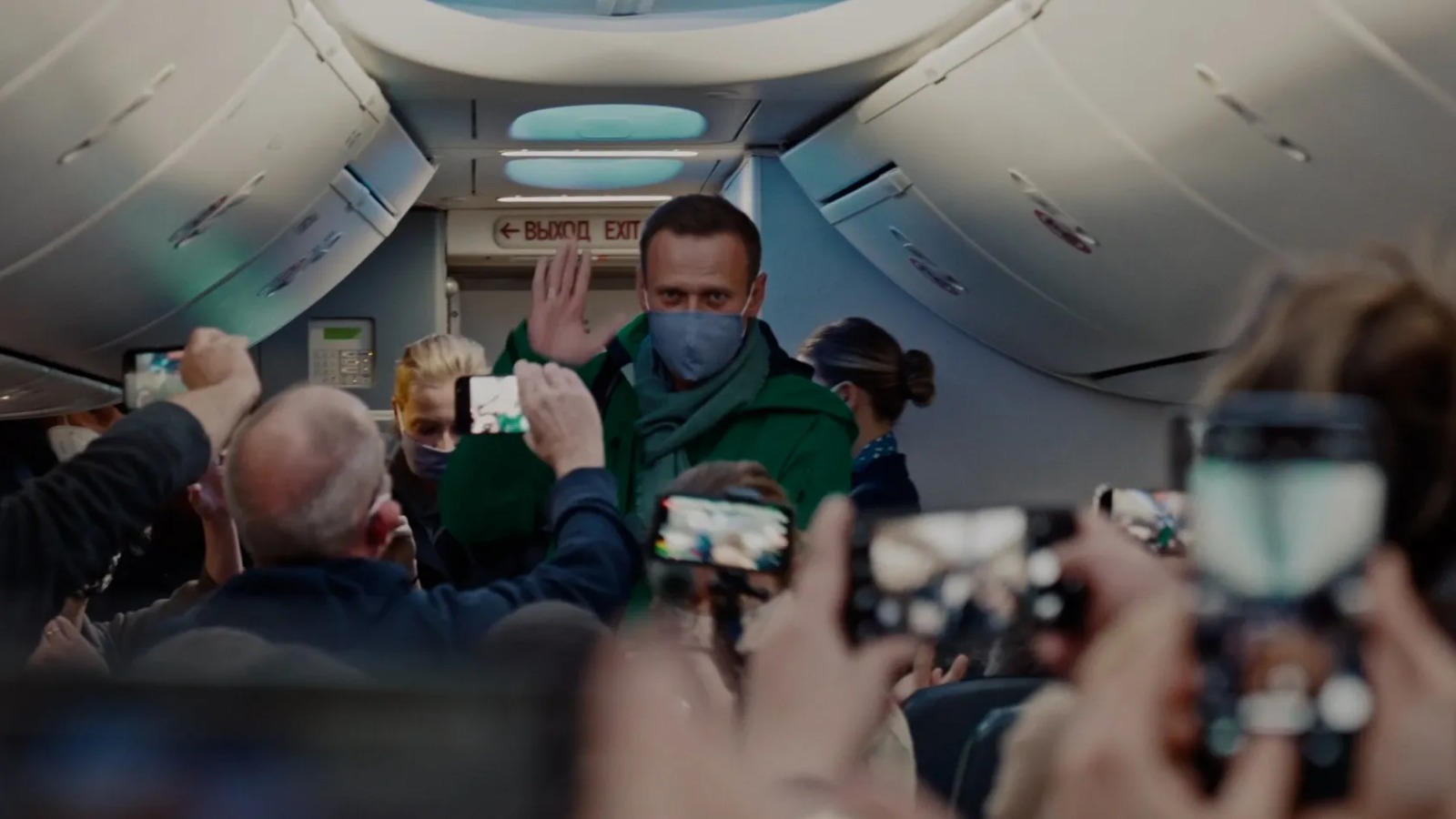 Cena do documentário Navalny. Na cena, vemos, ao fundo, Alexey Navalny, um homem branco, aparentando ter cerca de 40 anos, de cabelos castanhos curtos, usando máscara, cachecol e um casaco verde com a mão direita em um aceno, em um avião. A frente dele, vemos pessoas de costas com celulares filmando em sua direção.