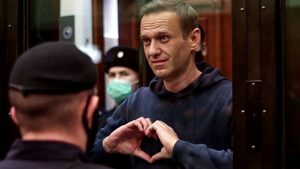  Cena do documentário Navalny. Na cena, vemos Alexey Navalny, um homem branco, aparentando ter cerca de 40 anos, de cabelos castanhos curtos, por detrás de um vidro, fazendo um símbolo de coração com as duas mãos à frente do peito. Ao seu lado e a sua frente, vemos oficiais de segurança usando máscaras.