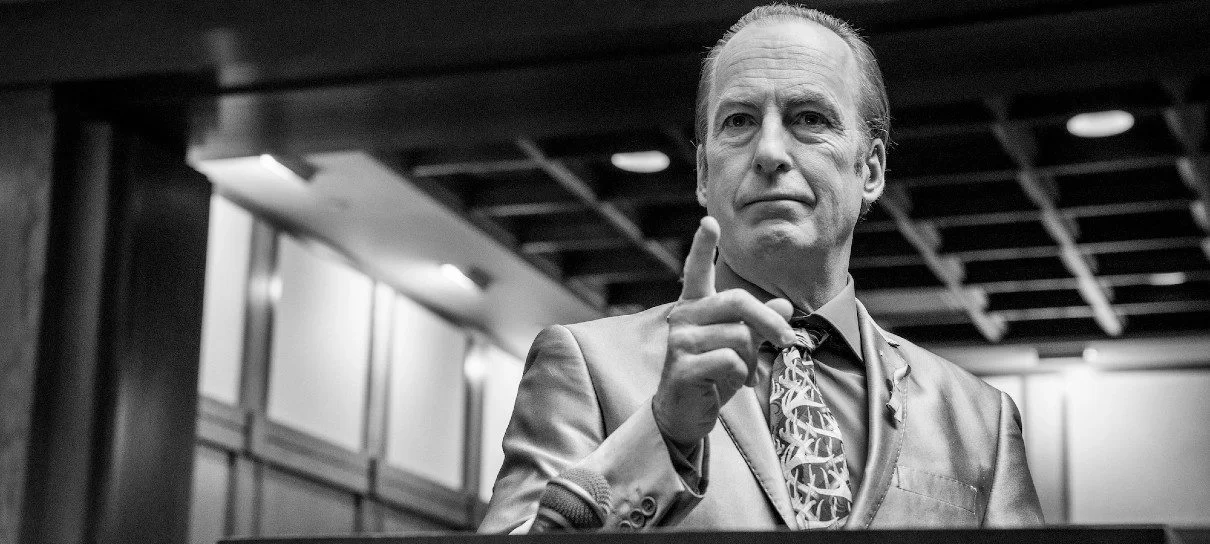 A foto é uma cena em preto e branco da sexta temporada de Better Call Saul, mostrando o protagonista Saul Goodman, interpretado por Bob Odenkirk, durante seu julgamento. O personagem está em pé, usando um terno e gesticulando com a mão.