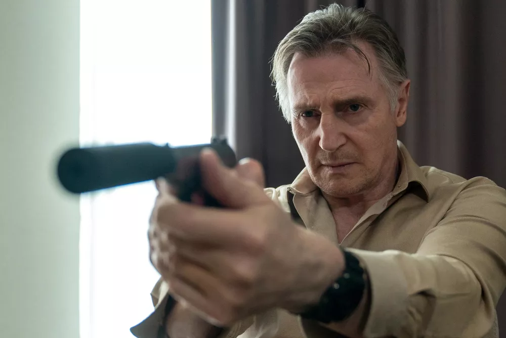 Cena do filme Assassino Sem Rastro, Alex, (Liam Neeson), homem branco de cabelos grisalhos, veste um casaco marrom claro e um relógio preto, aponta uma pistola com um silenciador na ponta