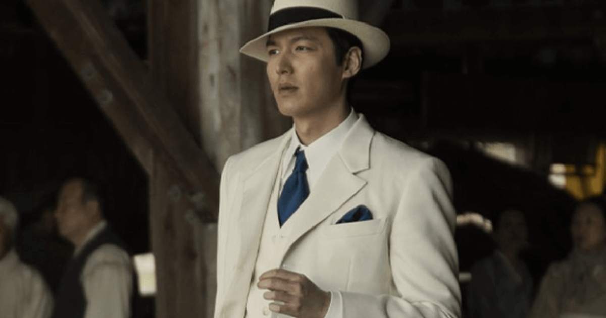 cena da série Pachinko (2022), Koh Hansu (Lee Min-ho) de pé em um salão, usando um terno branco, uma gravata azul e um chapéu com uma fita preta