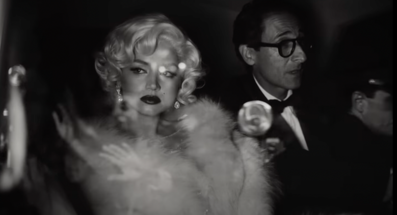 Cena do filme Blonde. Na imagem, Ana de Armas, que interpreta Marilyn Monroe, é uma mulher branca e loira. Ela está dentro de um carro ao lado de Arthur Miller, interpretado por Adrien Brody, um homem branco mais velho que usa um terno e um óculos.