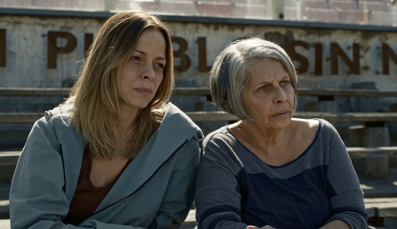 Cena do filme Ana. Sem Título. Na imagem temos, à esquerda, a atriz brasileira Stella Rabello. Ela tem cabelos loiros um pouco acima do busto, usa uma jaqueta na cor cinza e uma blusa vermelha. Do lado direito, temos a cineasta Lúcia Murat. Ela tem cabelos grisalhos e curtos e usa uma blusa de manga longa azul. O cenário da foto é em uma arquibancada à luz do dia. Ambas estão com expressões concentradas, com olhares atentos ao horizonte.