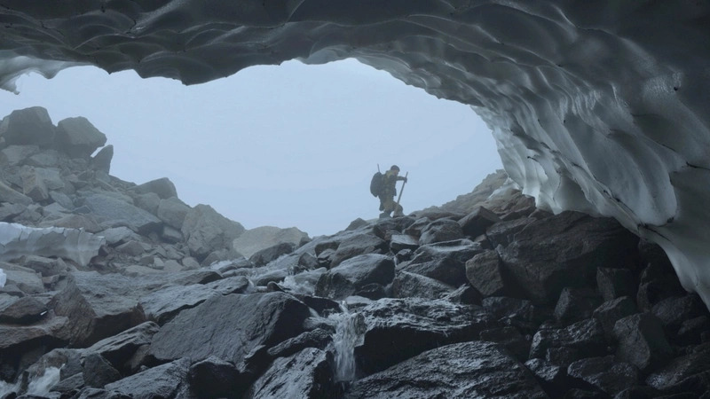 Cena do curta-metragem Haulout. Na imagem está o biólogo Maxim Chakilev. Ele aparece andando do lado de fora de uma caverna formada por grandes rochas cinzas. Ao fundo, o céu está tomado por neblina