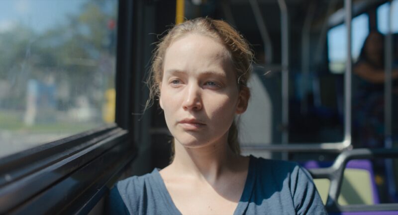 Cena do filme Causeway, da A24. Imagem retangular e colorida. Nela, a personagem Lynsey, interpretada por Jennifer Lawrence, está sentada no banco de um ônibus e olha contemplativa pela janela, com a luz do sol batendo contra seu rosto. Ela é uma mulher branca de cabelos loiros e olhos claros, que veste uma camiseta azul-escura.