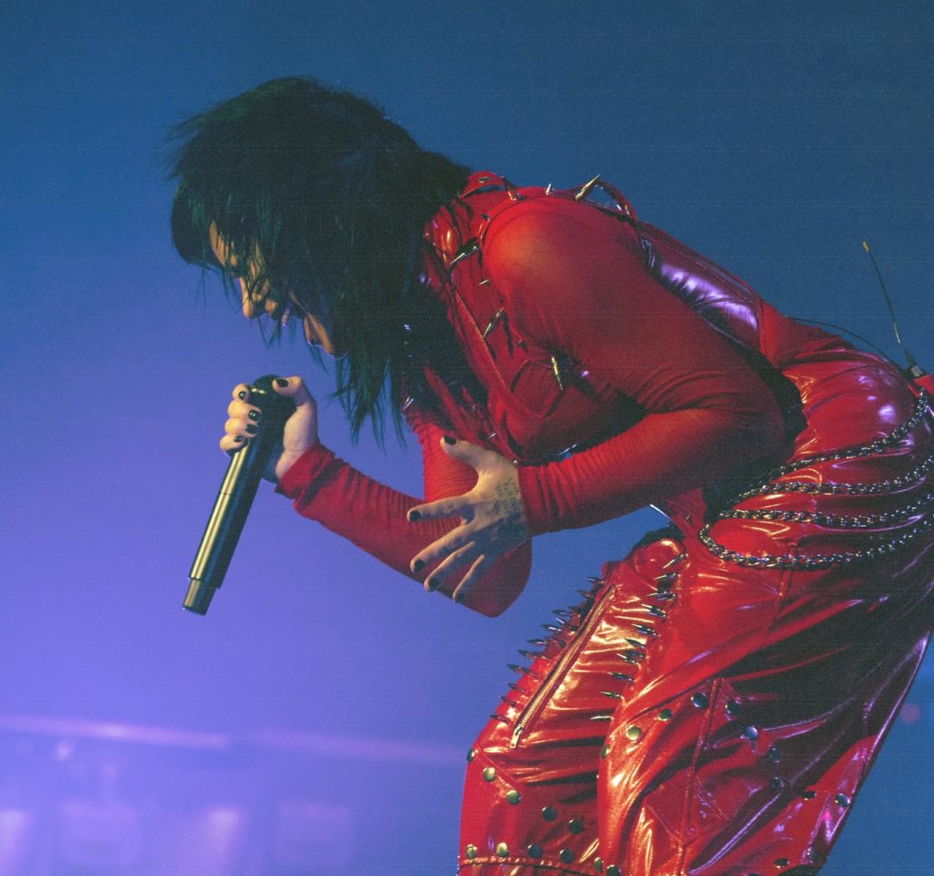 Foto de Demi Lovato no primeiro show da Holy Fvck Tour. A foto tem formato retangular, e mostra Demi com o microfone em uma das mãos, ela está em um movimento expressivo com o corpo enquanto canta com os cabelos ao vento.