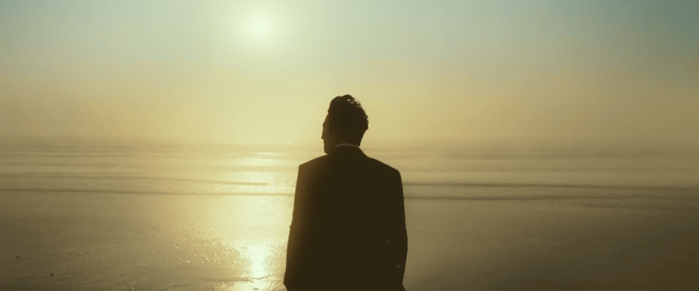 Cena do filme Decisão de Partir. Na imagem animada, Jang Hae-joon contempla o horizonte. A câmera captura as suas costas a partir da cintura. Jang é um homem sul-coreano de cabelos escuros. Ele veste um terno preto. Ao fundo da composição e a frente do protagonista, uma praia tomada pelo mar reflete o sol em suas ondas.