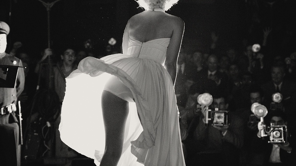 Cena do filme Blonde. A imagem mostra as costas da personagem Marilyn Monroe com a parte de baixo de seu vestido levantada, em uma interpretação de sua icônica cena no filme O Pecado Mora ao Lado. Imagem em preto e branco. 