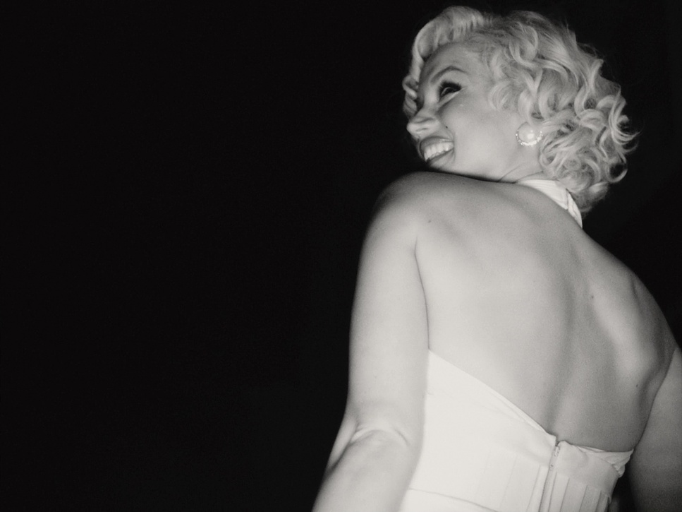 Cena do filme Blonde. Marilyn de costas, do tronco para cima, olhando para trás por cima do ombro e sorrindo. Em seu vestido branco para O Pecado Mora ao Lado. Foto em preto e branco