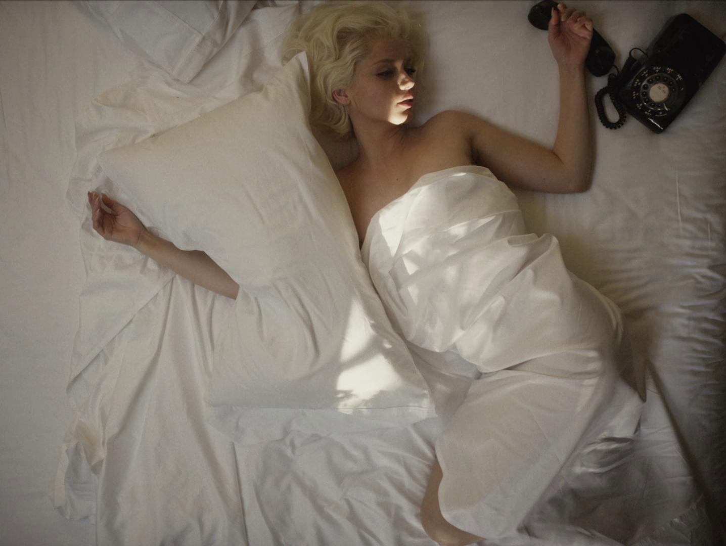 Cena do filme Blonde. Marilyn, já sem vida, deitada em sua cama, sem roupa, enrolada no lençol branco e com um travesseiro sobre seu braço direito. De seu lado esquerdo, um telefone desconectado, debaixo da sua mão
