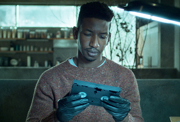 Cena da série Arquivo 81. Homem de cabelos escuros segura uma fita VHS preta na mão, enquanto a encara. Ele usa luvas pretas para segurar a fita e veste um suéter alaranjado.