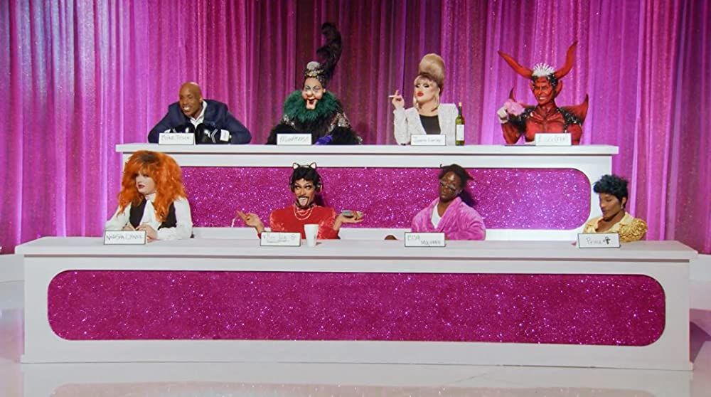 Cena da 7ª temporada de RuPaul's Drag Race: All Stars em que as oito participantes estão performando no Snatch Game. Há um palco com quatro queens em baixo e outras quatro em cima. Ao fundo há uma cortina rosa brilhante.