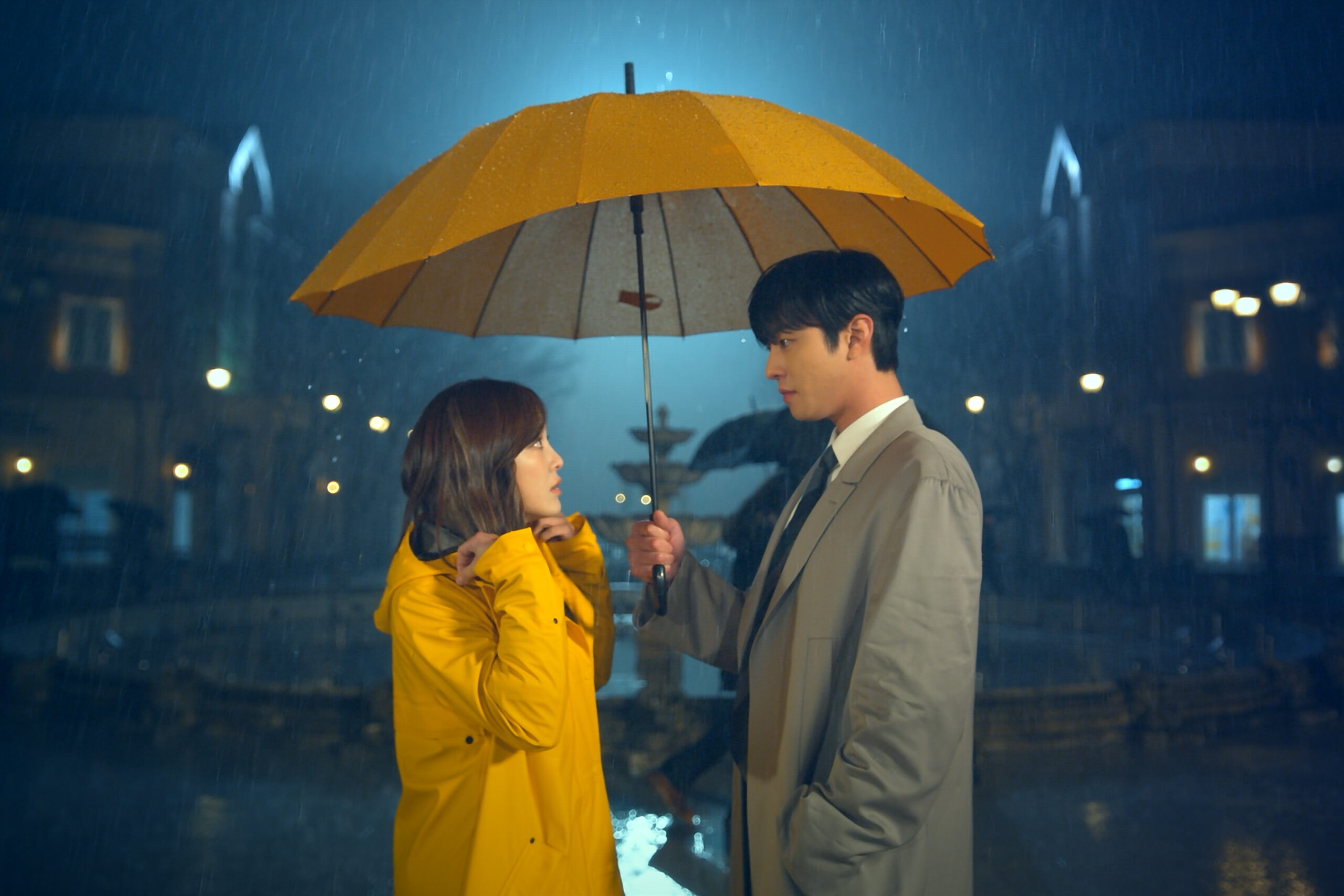 Cena da minissérie Pretendente Surpresa. Ha-ri (Kim Se-jeong) e Tae-moo (Ahn Hyo-seop) estão parados, sob a chuva, encarando um ao outro. Ha-ri, à esquerda, é uma mulher coreana magra de cabelos castanhos-escuros longos, usando uma capa de chuva amarela que cobre o seu corpo. Tae-moo, à direita, é um homem coreano magro de cabelos escuros e curtos, usando um sobretudo cinza-claro por cima de um terno social. Ele segura um guarda-chuva amarelo na mão direita, protegendo ele e Ha-ri da chuva. Atrás deles, podemos ver uma praça iluminada pela luz da lua e as silhuetas de vários prédios, contornadas pela luz de postes. Os dois estão centralizados na imagem, perdidos no olhar um do outro.