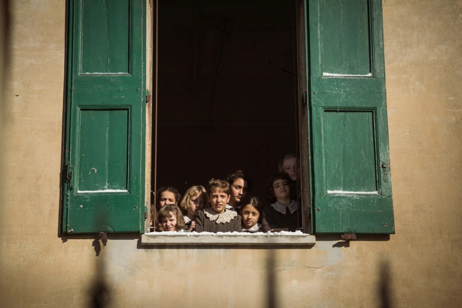 A foto mostra uma cena do curta em que várias das meninas do orfanato estão na janela. A janela é verde escuro, a parede é bege e as meninas estão usando roupas cinzas