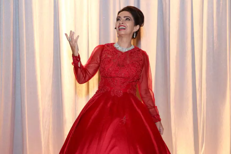 Cláudia Ohana, trajada com o clássico vestido vermelho de Maria Callas, sorri enquanto olha para cima, levantando sua mão direita em poética interpretação em frente a um fundo com cortinas brancas reluzentes