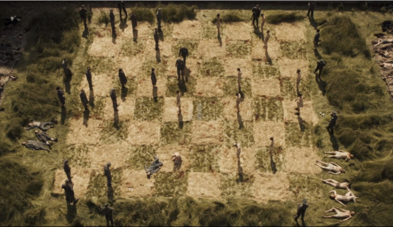 Texto Alternativo: Cena da série Hunters. A imagem apresenta um tabuleiro de xadrez gigante, em que suas divisões são a grama alta e a grama cortada. Suas peças são representadas por pessoas reais.
