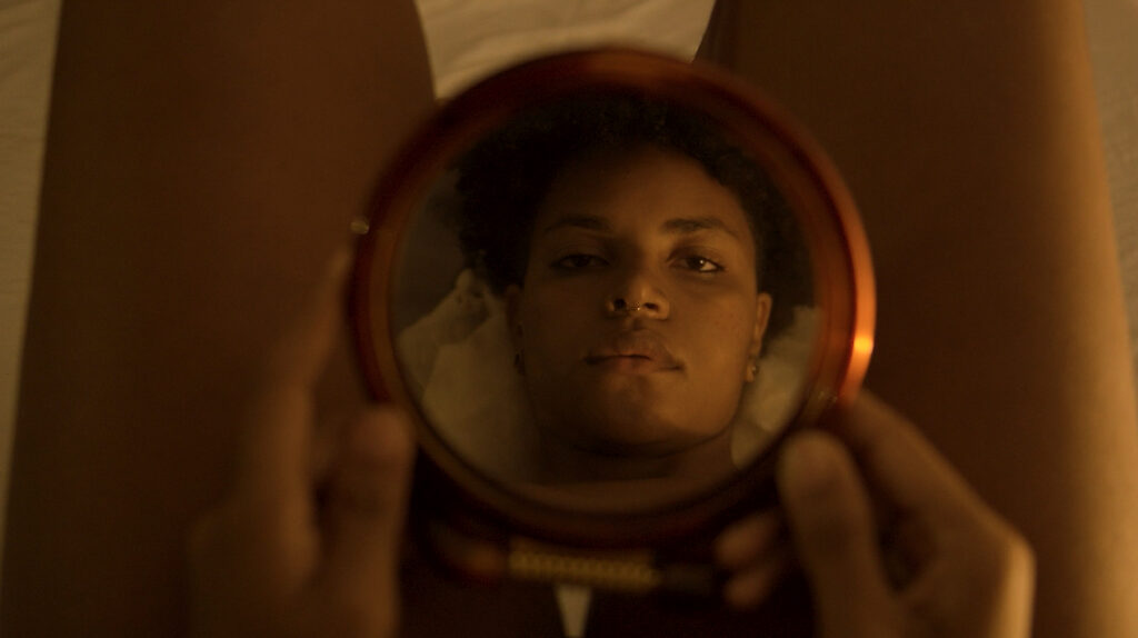 Cena do curta-metragem Todavia Sinto. Na imagem, a protagonista olha o seu reflexo em um espelho. Ela é uma mulher negra de cabelos crespos presos e segura o espelho entre as pernas.