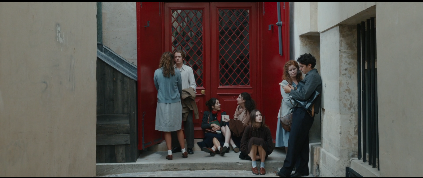 Cena do filme A Garota Radiante. Na imagem, sete jovens aparecem, na frente de uma porta vermelha: quatro estão de pé, e três estão sentadas.
