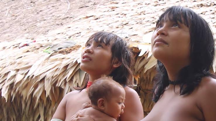 Cena do documentário Mãri hi - A Árvore do Sonho. Na imagem, duas mulheres indígenas de cabelos e olhos escuros parecem observar algo no céu. Uma delas segura uma criança no colo. A luz do Sol ainda está iluminando a paisagem. Ao fundo, o cenário é a aldeia onde vivem.