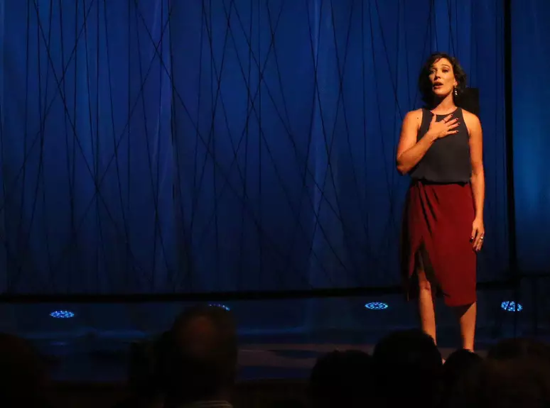 Cena da peça teatral Não!. A atriz Adriana Birolli está em primeiro plano, de pé, vestindo uma camisa preta com uma saia vermelha. No fundo do palco, há um quadro de fios pretos emaranhados em frente a uma enorme cortina branca, que ganha a cor azul através dos refletores