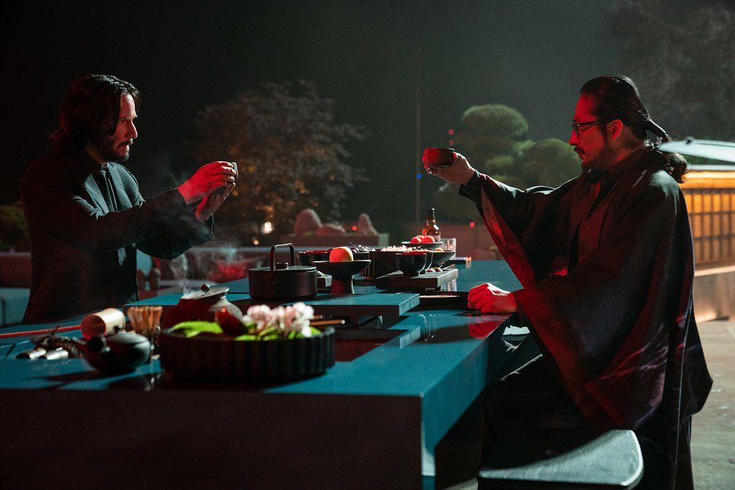 Dois homens sentados em uma mesa de frente para o outro. Ambos estão segurando xícaras de chá acima de seus cotovelos em um comprimento. O ambiente é marcado por um cenário rodeado por árvores em um templo japonês à noite. Iluminados por uma luz vermelha.