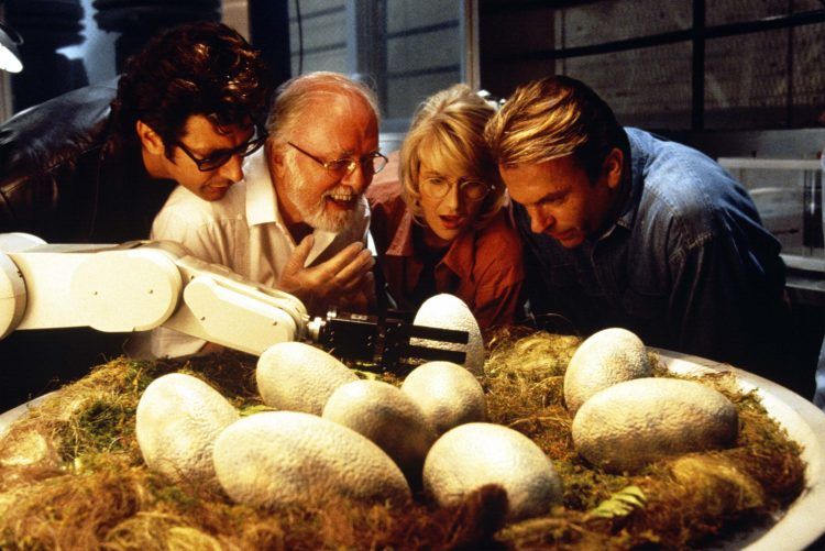 Cena do filme Jurassic Park. A foto mostra o elenco principal observando os ovos de dinossauro. Da esquerda para a direita. Ian, de casco preto e óculos escuros. John, de camisa branca e óculos de grau. Ellie, de camisa rosa e óvulos de grau. E Alan, de camisa jeans. Todos fascinados por nove ovos de dinossauro em um grande ninho. Uma mão robótica segura um dos ovos.