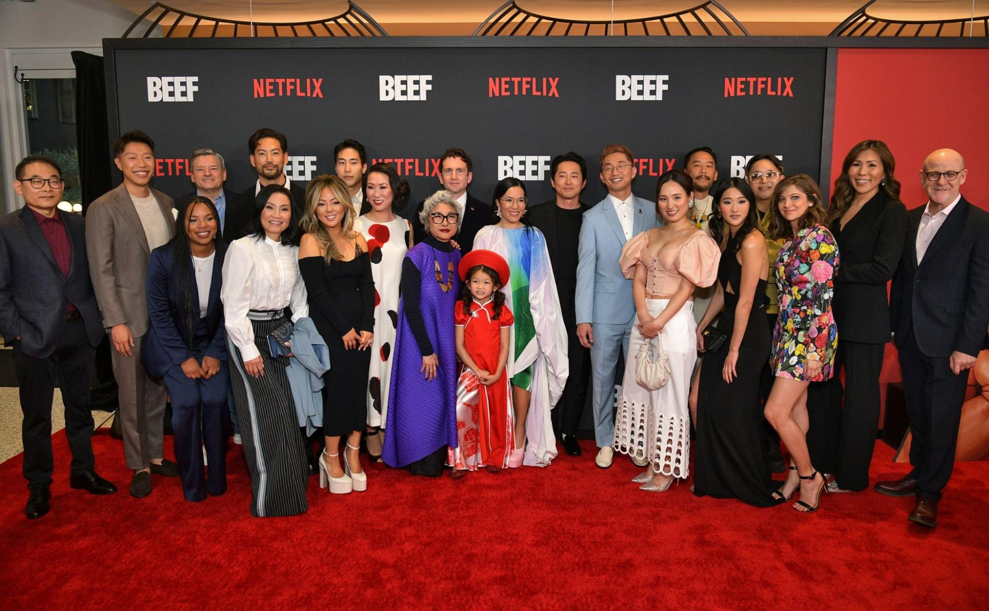  Foto da equipe da série Treta. Todos estão com roupas de gala em frente a uma parede com vários escritos repetidos das palavras “Netflix” e “Beef”. Todas as vinte e duas pessoas estão acima de um tapete vermelho e são majoritariamente asiático-americanos.
