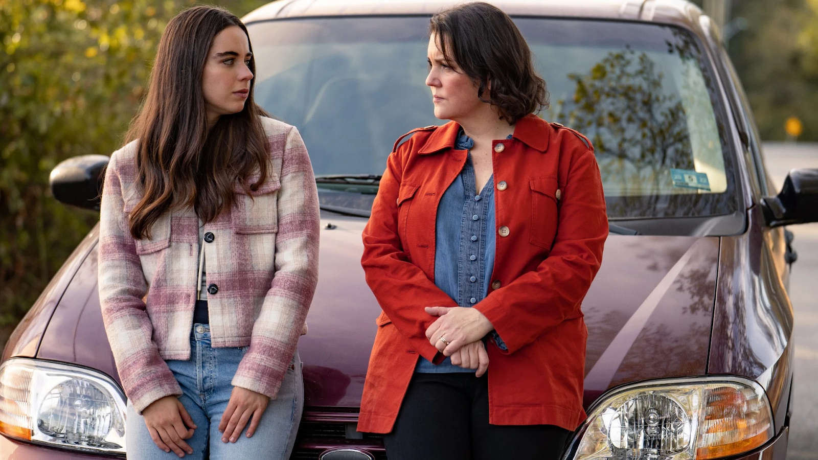 Cena da segunda temporada de Yellowjackets. Shauna (Melanie Lynskey) e Callie (Sarah Desjardins) conversam apoiadas no capô de um sedan vermelho. Shauna, à direita, é uma mulher caucasiana de meia idade, cabelos pretos até os ombros, usando uma jaqueta vermelha por cima de uma camisa azul e calças azuis-escuras. Callie, à esquerda, é uma adolescente caucasiana de cabelos castanhos-escuros longos, usando um casaco xadrez branco e rosa fechado com calças jeans. Shauna segura suas mãos, observando intensamente Callie, que por sua vez apoia suas mãos nas coxas, olhando temerosa para a mãe. As duas estão paradas no meio de uma estrada, durante o dia, e podemos ver alguns arbustos fora de foco no canto esquerdo da tela, enquanto a estrada continua pelo canto direito, também fora de foco.