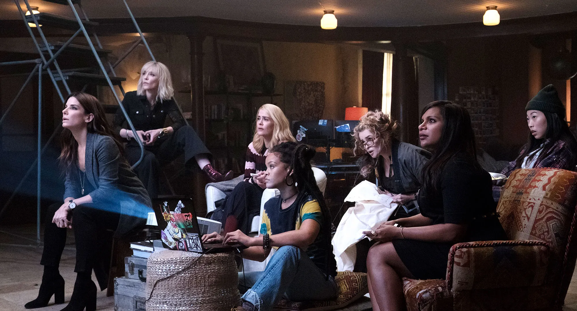 Cena do filme Oito Mulheres e um Segredo. As atrizes encontram-se sentadas em um galpão escuro, analisando a projeção do plano para roubar o colar.
