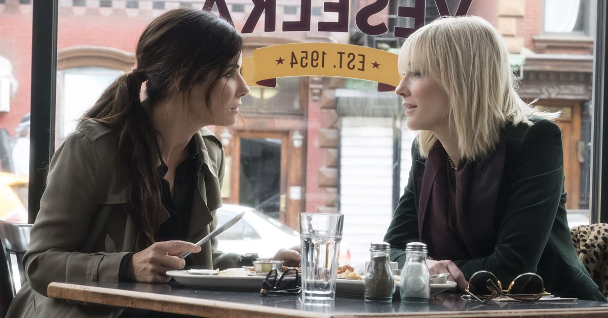 Cena do filme Oito Mulheres e um Segredo. Sandra Bullock e Cate Blanchett sentadas em uma mesa com dois pratos com comida, em um café conversando.