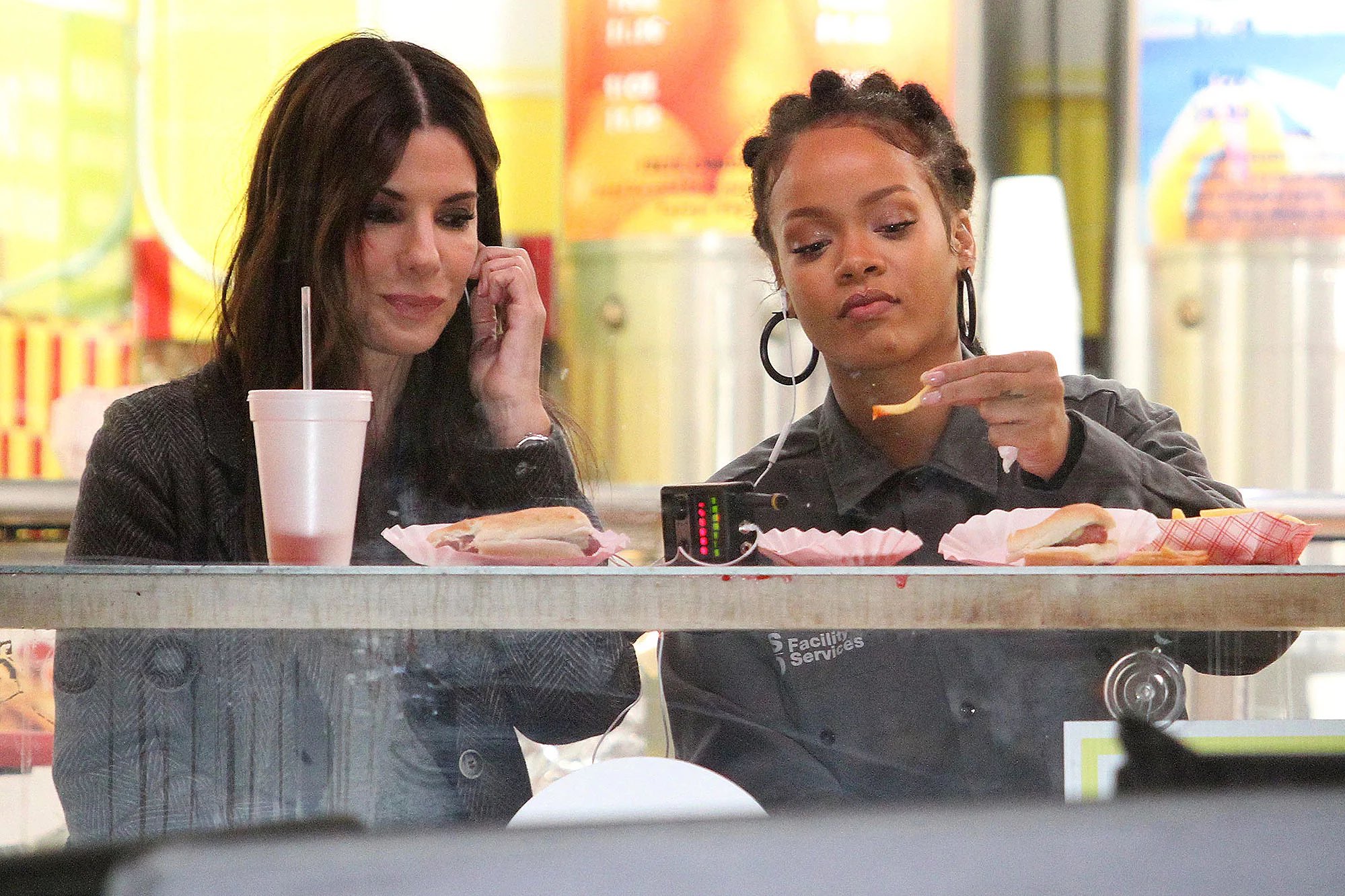 Cena do filme Oito Mulheres e um Segredo. Sandra Bullock e Rihanna compartilhando um fone de ouvido comendo batata frita e cachorro-quente em uma lanchonete.
