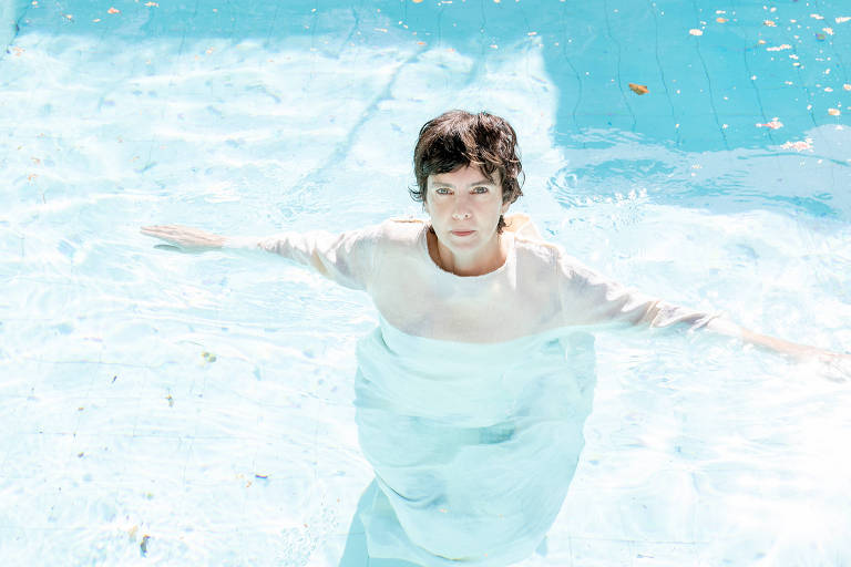 A artista está flutuando numa piscina azul, de vestido, olhando fixamente para a câmera.